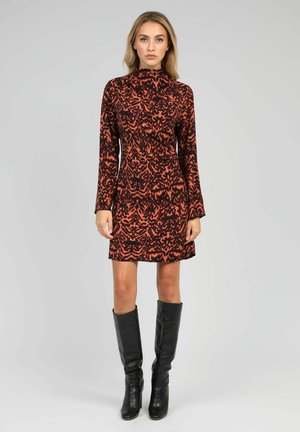 Femme portant une robe à manches longues et col montant, orange avec un motif noir, et des bottes en cuir noires montant jusqu'aux genoux, debout devant un fond uni.