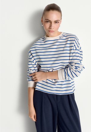 Femme aux cheveux tirés en arrière portant des boucles d'oreilles créoles argentées, une chemise blanche à manches longues avec des rayures bleues, et un pantalon sombre, debout contre un mur blanc.