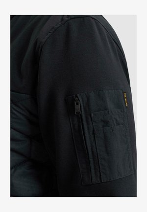 Schwarzer Jackenärmel mit einer Reißverschlusstasche und kleineren aufgenähten Fächern, mit einem kleinen gelben „PME Legend“ Stoffetikett.