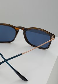 Óculos de sol em tartaruga com lentes azuis e detalhes em metal. Apresentam um design elegante e angular, com hastes em contraste de verde escuro e dourado.