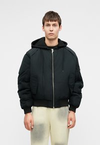 Fekete puff jacket kapucnival, elülső cipzárral és nagy oldalsó zsebekkel. Bordázott mandzsetták és derék. Sötét felsőn és világos nadrágon viselve.