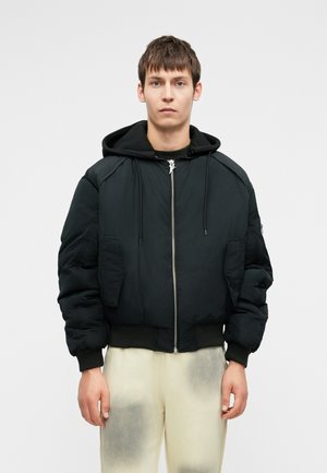 Crna puffer jakna s kapuljačom, prednjim zatvaračem i velikim bočnim džepovima. Pletivni manžeti i donji rub. Nosena preko tamne majice i svijetlih hlača.