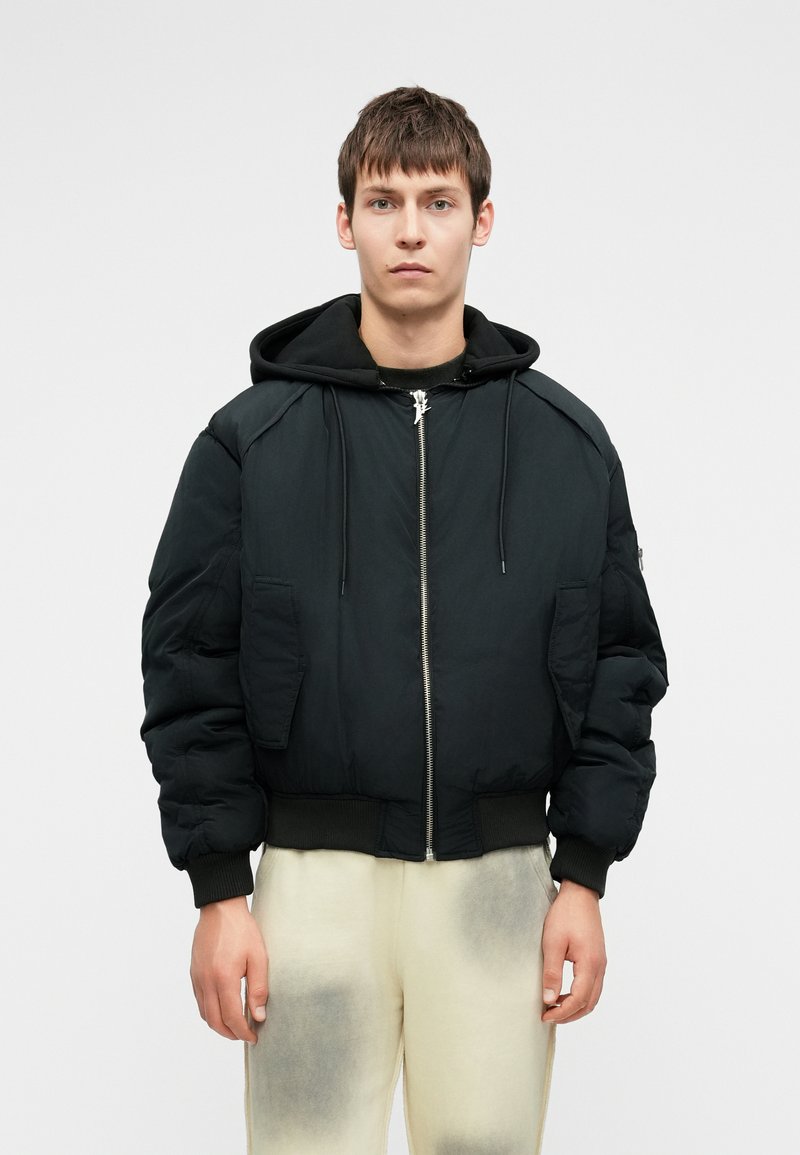 Fekete puff jacket kapucnival, elülső cipzárral és nagy oldalsó zsebekkel. Bordázott mandzsetták és derék. Sötét felsőn és világos nadrágon viselve.