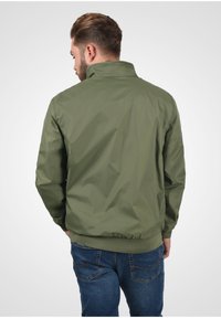 Olivenfarget, lett jakke med høy krage, ribbede mansjetter og nederkant, som har en glatt tekstur og minimalistisk design.