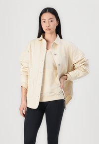 Veste en velours côtelé beige clair avec fermeture boutonnée, offrant une coupe décontractée, deux poches avant et une texture douce, portée sur un haut assorti et un legging noir.