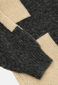 Pull en maille présentant un mélange de couleurs gris foncé et crème, avec un motif texturé et un détail de bord-côte aux poignets.