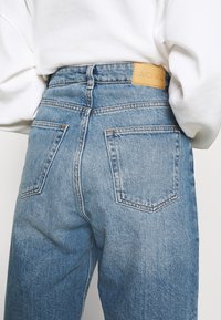 Högmidjade blå denimjeans med en brun läderetikett, två bakfickor och en texturerad tyg. Avslappnad passform med synliga söm detaljer.
