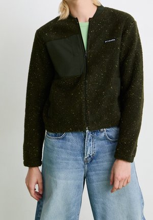 Person trägt eine dunkelgrüne Fleece-Jacke mit Reißverschluss, schwarzem Brusttaschen-Patch und hellblauen, hoch taillierten Jeans, steht vor einem neutralen Hintergrund.