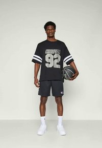 T-shirt preto de mangas curtas com gráfico "CHAMPION 92", combinado com calções escuros e ténis brancos. Ele segura uma bola de basquete preta.