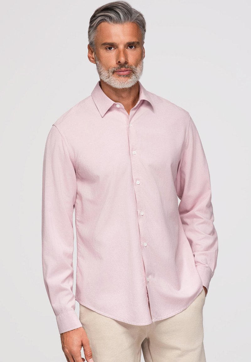 Homme aux cheveux gris et à la barbe, portant une chemise rose clair à boutons et un pantalon beige, debout devant un fond clair uni.