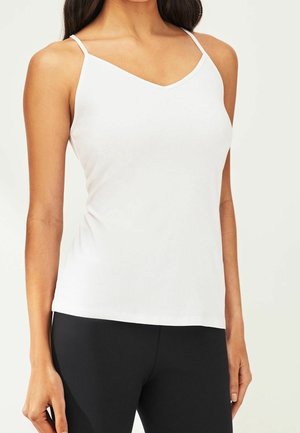 Kobieta ubrana w biały, bezrękawnik z dekoltem w kształcie litery V i cienkimi ramiączkami oraz czarne dopasowane spodnie na jednolitym tle.