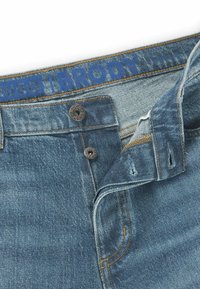 Vaaleansiniset farkut, joissa on nappikiinnitys, kontrastivärinen ompelu ja "HUGO BRODY" -logo painettuna vyötärönauhaan.