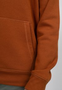 Roodbruine sweatshirt met een voorzak, ribgebreide mouwen en onderkant, met zichtbare stikdetails. Zachte textuur, casual ontwerp.