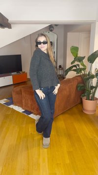 Szürke kötött pulóver, sötétkék farmer, és világos bézs bokacsizma. A szobában fa padló, barna kanapé és szobanövények találhatók.