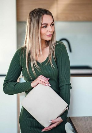 Beige kožená kabelka s embosovaným logem, obdélníkový tvar, držena jednou rukou. Pozadí tvoří světlé dřevo a minimalistická dekorace.