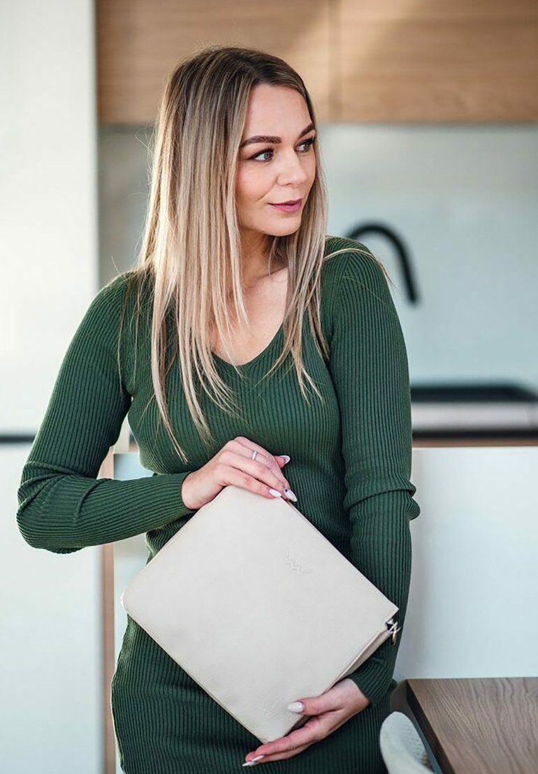 Beige kožená kabelka s embosovaným logem, obdélníkový tvar, držena jednou rukou. Pozadí tvoří světlé dřevo a minimalistická dekorace.