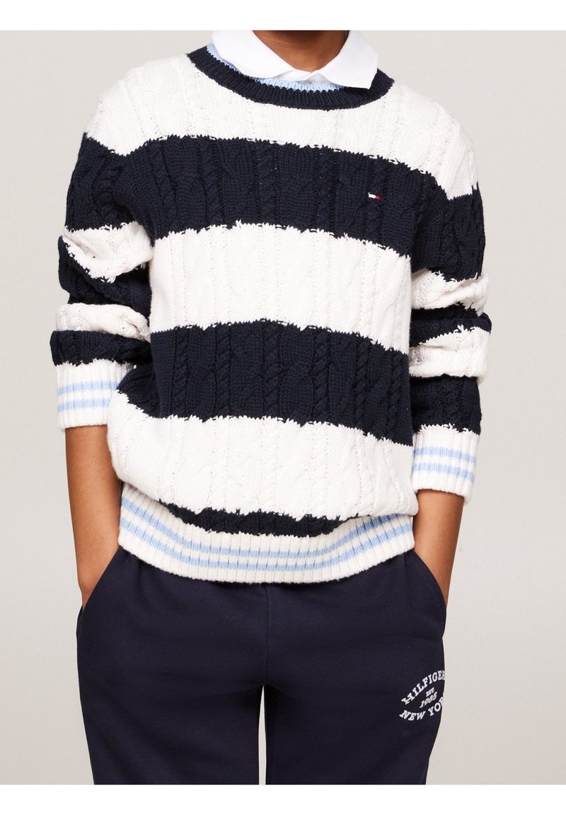 Personne portant un pull en maille torsadée rayé bleu marine et blanc par-dessus une chemise blanche à col, avec un pantalon de survêtement bleu marine avec un texte blanc sur la jambe gauche.