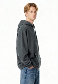 Sötétszürke kapucnis pulóver kenguruzsebbekkel, húzózsinórral, laza fazonnal és bordázott mandzsettákkal. Kék farmernadrággal párosítva, világos háttér előtt állva.