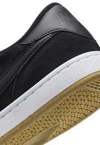 Mustat mokka- ja nahkakenkä, jossa valkoinen kumipohja. Huomattavia teksturoituja yksityiskohtia ja kohotettu logo kantapäällä.