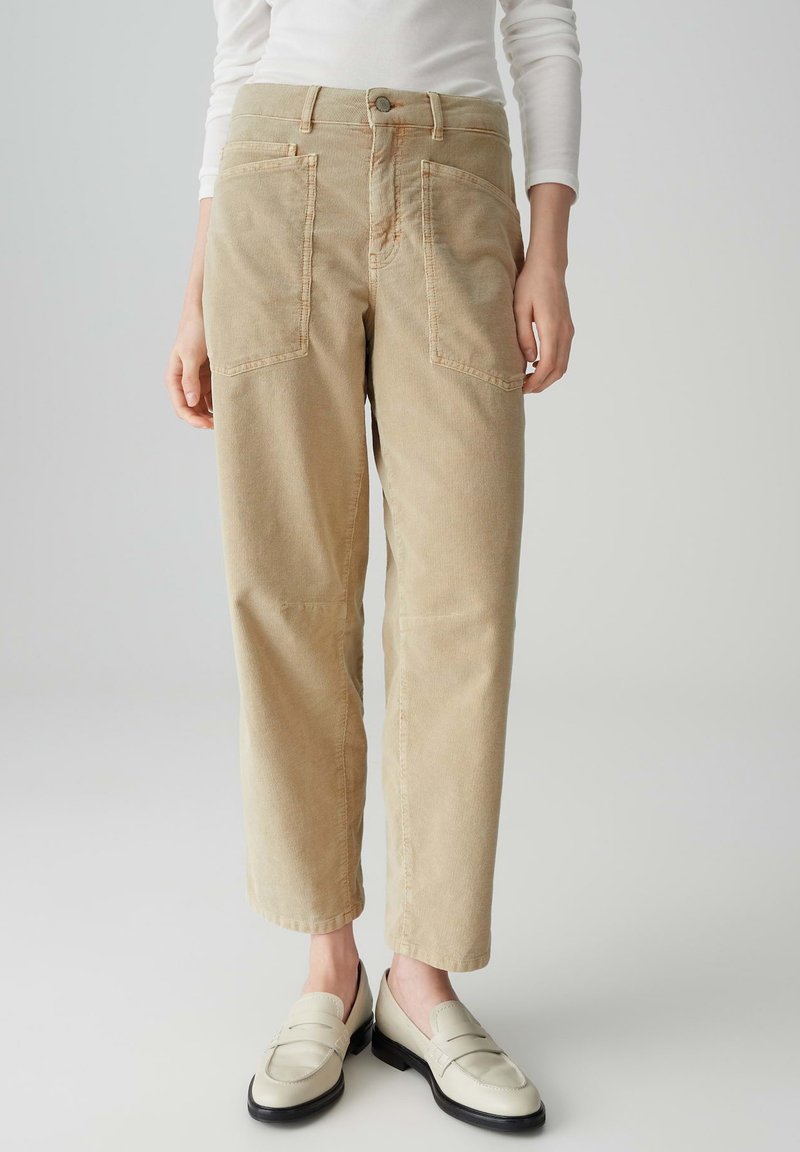 Beige corduroy broek met wijde pijpen, voorzien van twee voorzakken en een knoopsluiting bij de taille. Draagt met lichtgekleurde instappers.