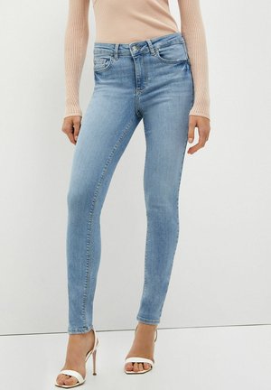Jeans skinny azzurro chiaro abbinati a una maglia beige a coste a maniche lunghe e sandali bianchi con tacco alto e punta aperta.