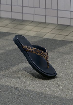 Infradito nera con cinturino in tessuto con stampa leopardata che fluttua sopra l'asfalto vicino a un muro piastrellato.