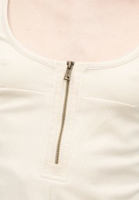 Κοντινή λήψη λευκού ριμπ ύφανσης τοπ με μεταλλικό φερμουάρ στο μπροστινό μέρος που είναι μερικώς κλειστό.