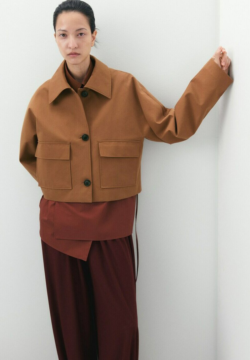 Femme portant une veste courte marron avec de grandes poches et une tenue superposée rouge foncé, debout avec un bras appuyé contre un mur blanc.