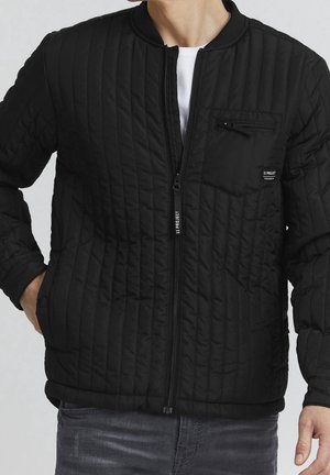 Hombre con chaqueta acolchada negra con bolsillo en el pecho con cremallera y cremallera frontal, combinado con vaqueros oscuros y una camisa blanca debajo.