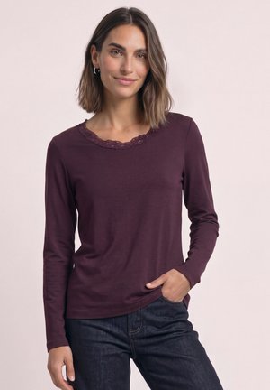 Kvinde med skulderlangt brunt hår iført en langærmet mørkelilla skjorte med blonder og mørke jeans, stående med den ene hånd i lommen.