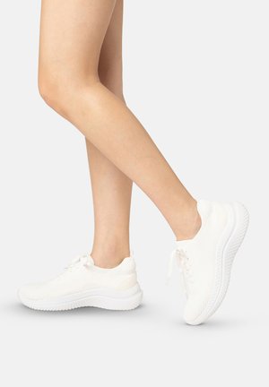 Benenden die witte gebreide sneakers met dikke zolen dragen, één voet plat en de andere op de tenen, tegen een egale lichte achtergrond.