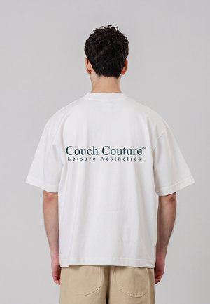 Mand med kort mørkt hår iført en hvid T-shirt med teksten ""Couch Couture Leisure Aesthetics"" trykt på ryggen.
