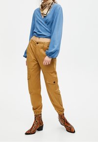 Blouse en tissu bleu à manches longues, pantalon cargo beige avec poches latérales, et bottines marron à imprimé serpent avec talon bloc.