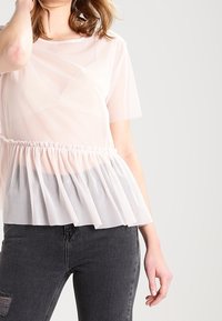 Světle růžové tričko s průhlednou vrstvou, krátkými rukávy a nařaseným tylovým lemem. Nošeno s tmavými džíny, má kulatý výstřih a jemnou texturu.