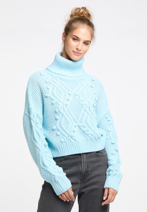 Lichtblauwe gebreide trui met een hoge geribbelde kraag, met textuurpatronen en pompondetails, gecombineerd met donkergrijze jeans.