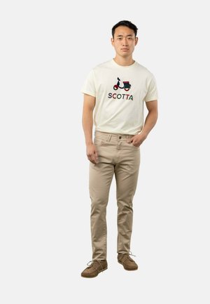 Hombre de pie con camiseta crema con gráfico de scooter y texto "SCOTTA", pantalones beige y zapatos marrones con cordones sobre fondo blanco.