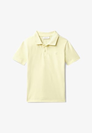 Junge steht mit einer Hand in der Tasche, trägt ein hellgelbes Poloshirt, schwarze Hose und schwarze Schuhe vor einem schlichten grauen Hintergrund.