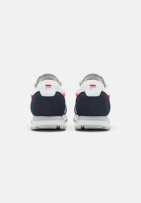 Hvite og marineblå sneakers med røde detaljer, laget av semsket skinn og mesh-materialer, rund form og teksturert gummisåle. Merkevarelogo i rødt på hælen.