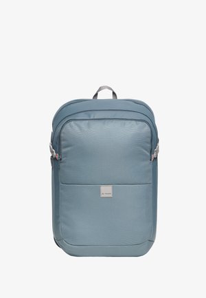 Mochila rectangular azul claro con bolsillo frontal con cremallera, asa superior, correas de compresión laterales y una pequeña etiqueta en el panel frontal.