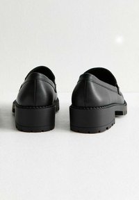 Scarpe in pelle nera con punta arrotondata, tacco basso a zeppa e dettaglio laterale testurizzato. La suola presenta scanalature orizzontali per una migliore aderenza.