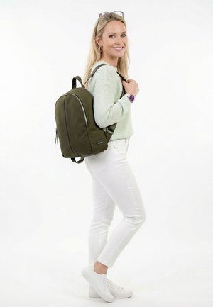 Mujer sonriente con suéter verde menta y pantalones blancos, llevando una mochila verde oliva sobre un hombro contra un fondo blanco.