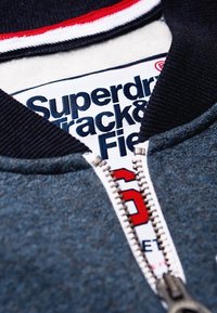 Blå zip-jacka med marinblå ribbad krage; har röda och vita accenter. Etiketten visar "Superdry Track & Field," med en mönstrad dragkedja.