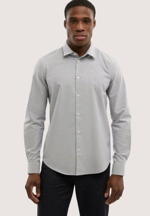 Mężczyzna w jasno szarej, długorękawowej koszuli z kołnierzykiem i guzikami, w ciemnych spodniach, stojący na jednolitym tle.