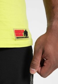 Neon geltonos spalvos marškiniai su stačiakampiu guminio ženklo užrašu "GORILLA WEAR UNITED STATES OF AMERICA" kairiajame šone. Matomi juodi šortai.