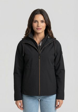 Femme aux longs cheveux bruns portant une veste noire zippée avec des cordons de serrage et un jean bleu clair, debout devant un fond clair uni.