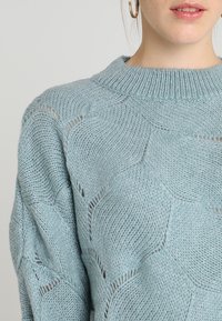 Jasnoniebieski sweter dziany z teksturowanym wzorem plastra miodu i okrągłym dekoltem. Materiał jest miękki z widocznymi detalami ściegów.