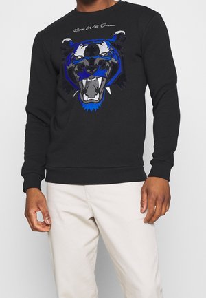 Schwarzer Sweatshirt mit einer blau-schwarzen Grafik eines brüllenden Tigers und weißem Text "Keep Well Dream", getragen zu hellbeigen Hosen.