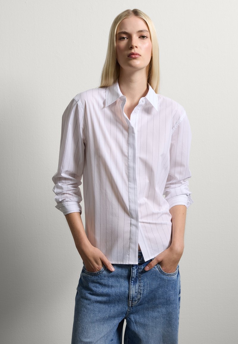 White button-up shirt with vertical light gray stripes, featuring a classic collar and rolled sleeves, paired with loose blue denim jeans.