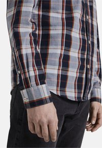 Chemise pour homme à manches longues avec boutons, présentant un motif à carreaux bleu marine, blanc et rouge, fabriquée en tissu léger à la texture lisse.