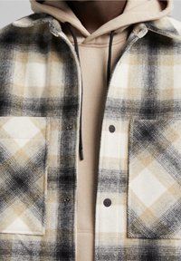 Persona con una sudadera beige debajo de una chaqueta de cuadros en negro, blanco y marrón con botones a presión y bolsillos en el pecho, mostrada de cuello a media torso.